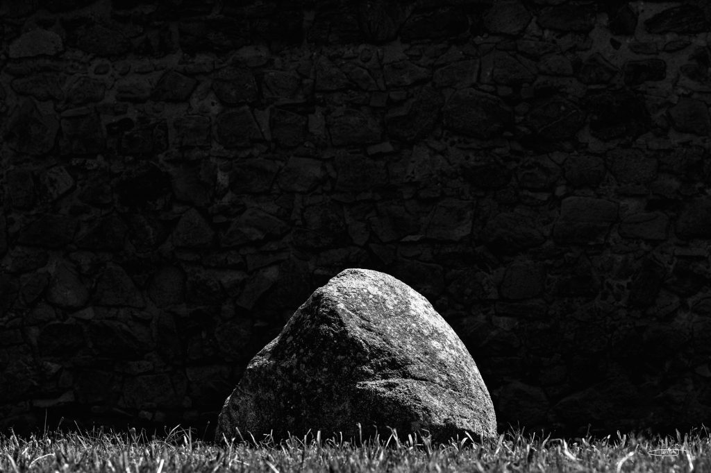 Pedra iluminada diante de um muro escuro, numa composição fotográfica a preto e branco.